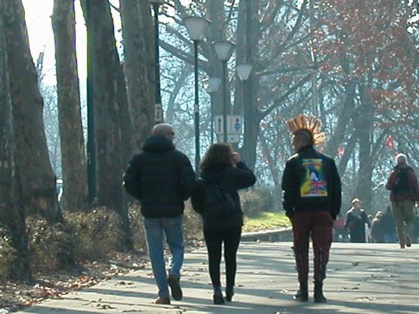 People strolling on tree-lined path in urban park, one with yellow mohawk