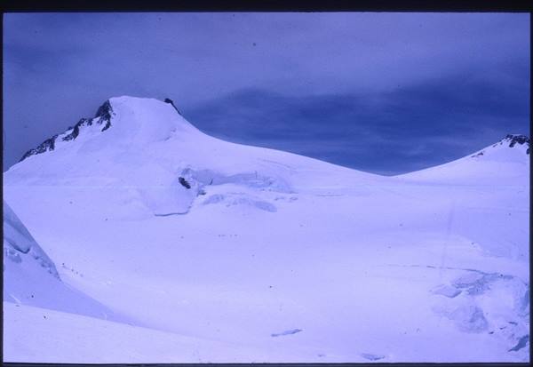 Snow-covered summit with crevasses and dark rocks under cloudy sky