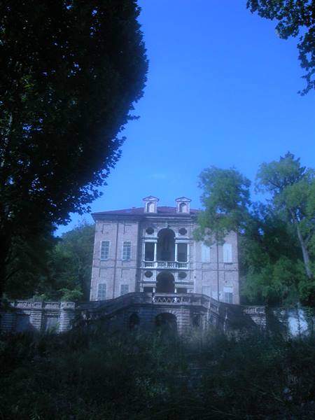 Abandoned historic villa with stone staircase, framed by trees and overgrown grass