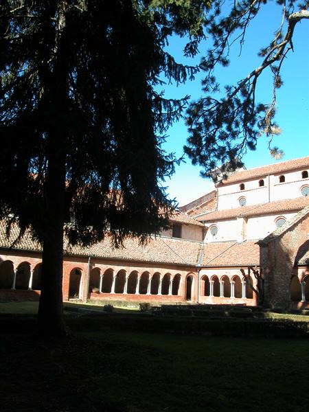 Brick cloister with arched arcade and abbey church, pine tree in foreground, sunlight
