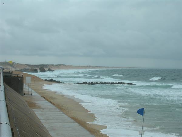 Waves on the beach under cloudy sky, blue flag and concrete walkway
