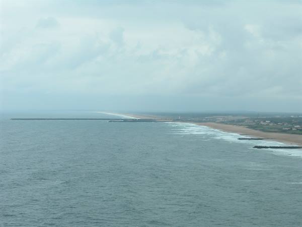 Ocean waves toward a sandy beach with pier and houses under cloudy sky