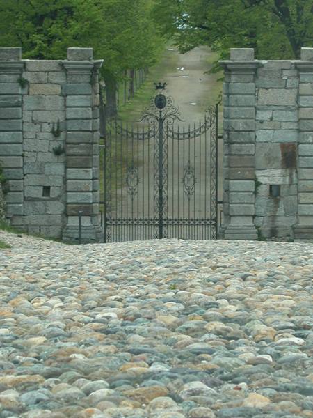 Wrought iron gate between grey stone pillars, tree-lined path beyond cobblestones