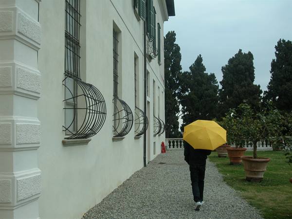 Person with yellow umbrella walking past white villa with curved iron window grilles, cypress trees behind