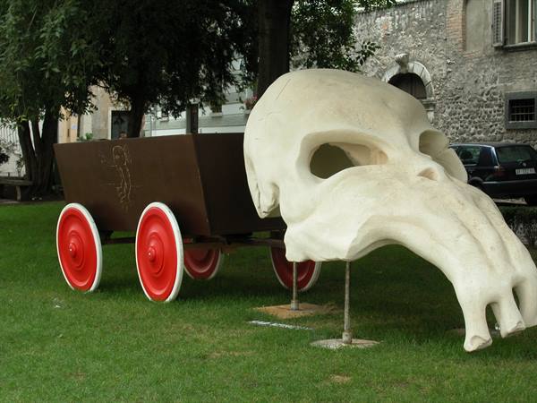 White skull sculpture with red-wheeled cart on an urban lawn