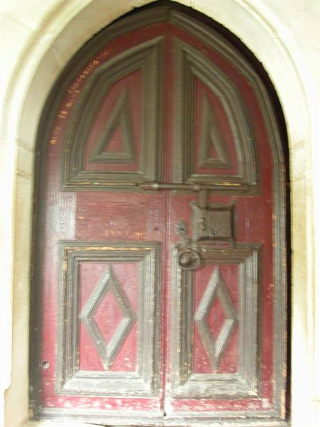 Red wooden door with carved diamonds and iron latch, framed by stone arch