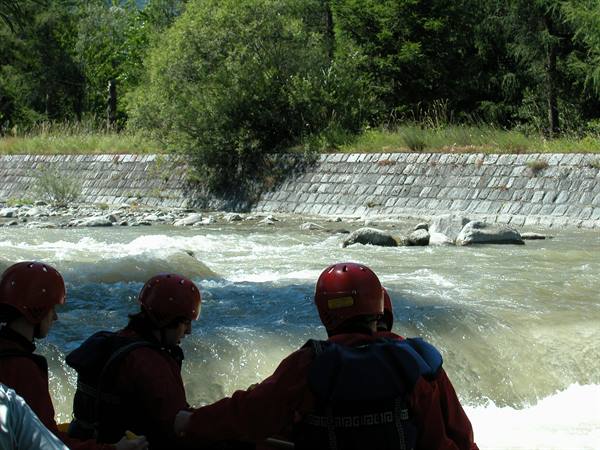 Rafters in red helmets ride the river past a stone embankment