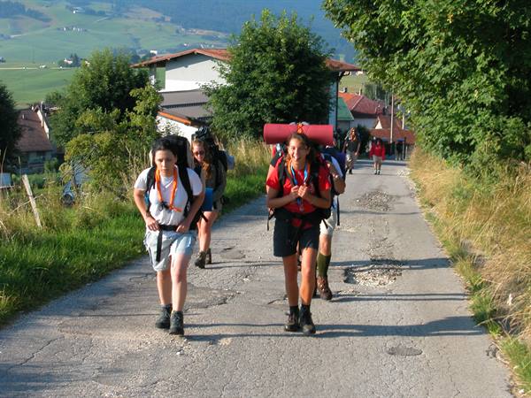 Young hikers with backpacks walk on an asphalt road past houses and green hills