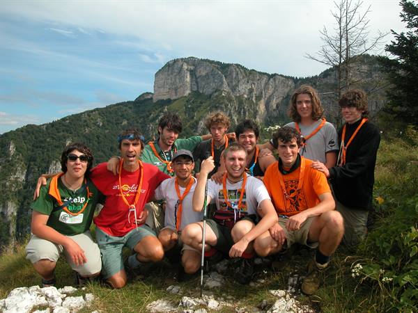 Group of young hikers with orange neckerchiefs crouching on mountain meadow below rocky cliff
