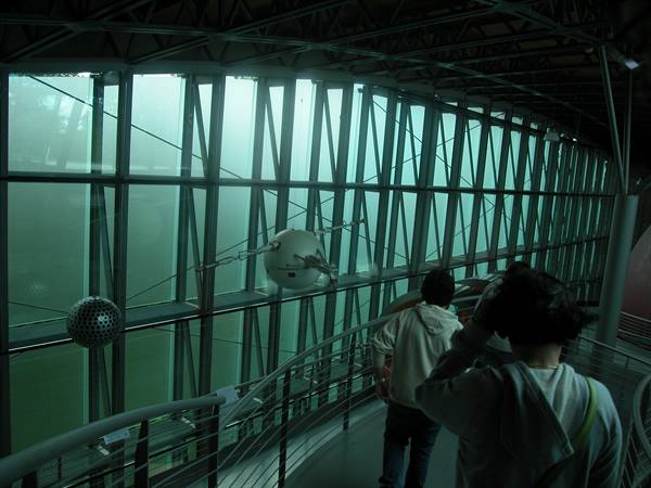 Steel ramp beside green glass wall, two visitors photographing sphere sculptures