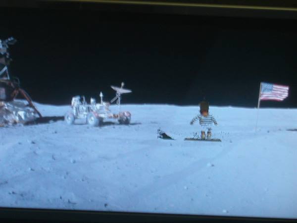 Screen showing lunar rover, American flag and small figure on gray surface