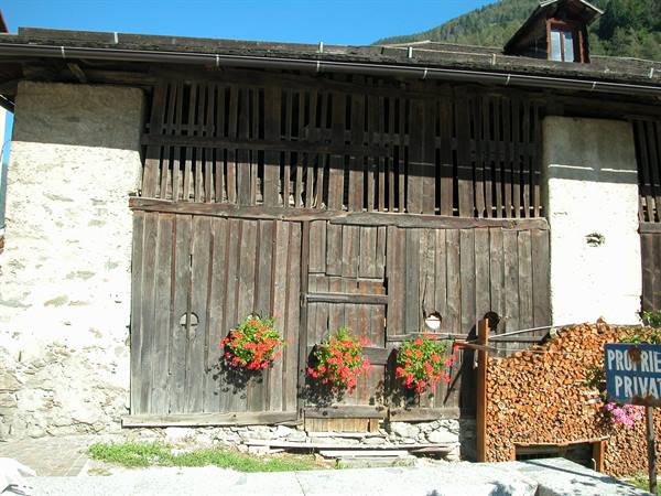 Wooden and stone facade with red hanging flowers, firewood stack, sunny mountain
