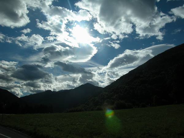 Sun through clouds above forested mountains and green meadow
