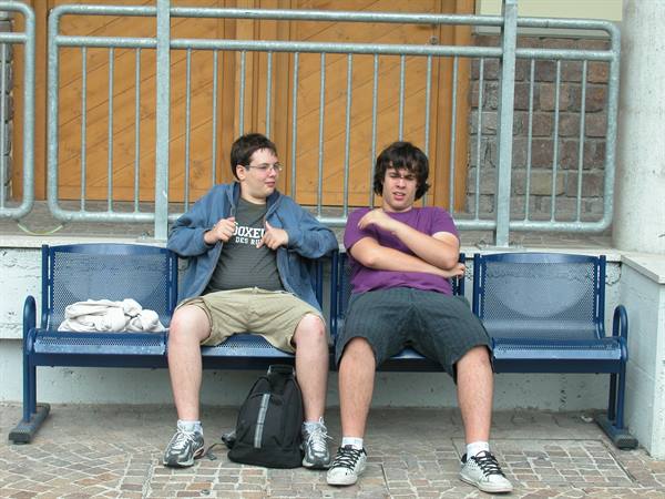 Two young men sit on a blue metal bench in front of a railing