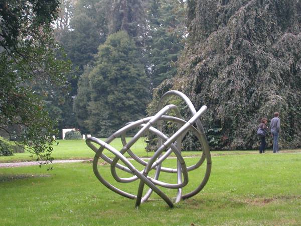 Sculpture of intertwined metal tubes on green lawn, visitors among trees