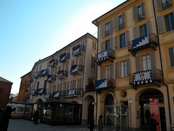 Ochre buildings with heraldic banners on balconies, arcades and pedestrians in sunlight