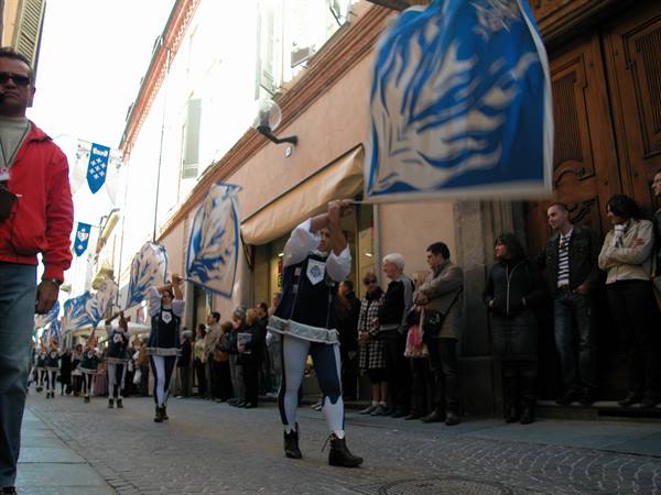 Flag wavers with blue banners parade through a narrow street, crowd watching