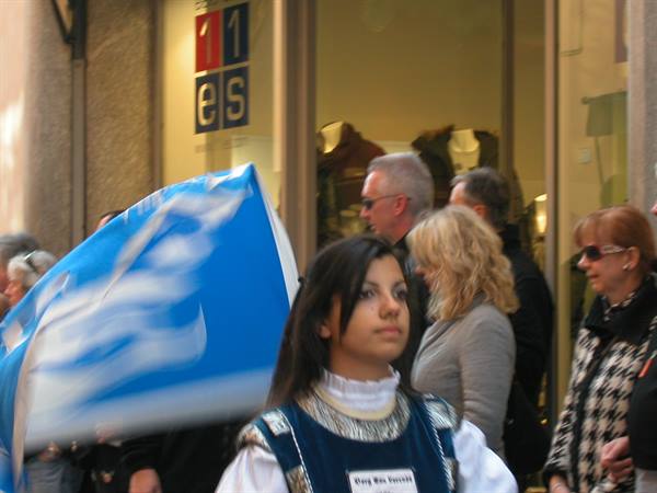 Girl in traditional costume with blue flag parading past a shop