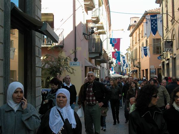 Crowd on Via P. Bretti, buildings with hanging flags, women in headscarves walking