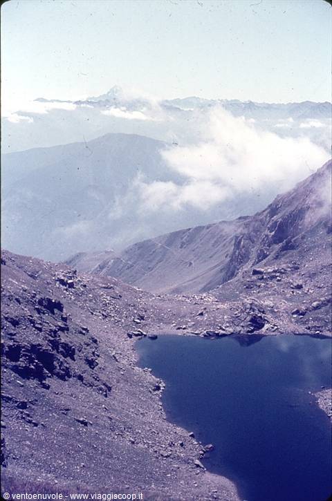 Alpine lake surrounded by rocky debris, low clouds on mountains, snow in distance