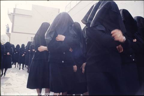 Women veiled in black marching between whitewashed houses, harsh daylight