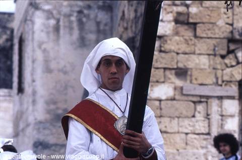 Young man in white robe and red sash carries a black cross by stone walls