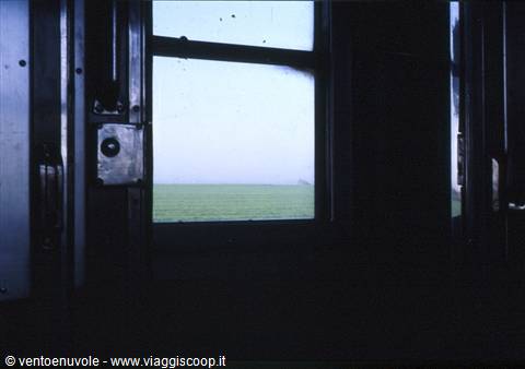 Train window framed by dark metal door, green fields in fog