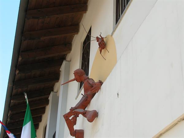 Iron sculpture of Pinocchio and cricket on white facade, wooden roof
