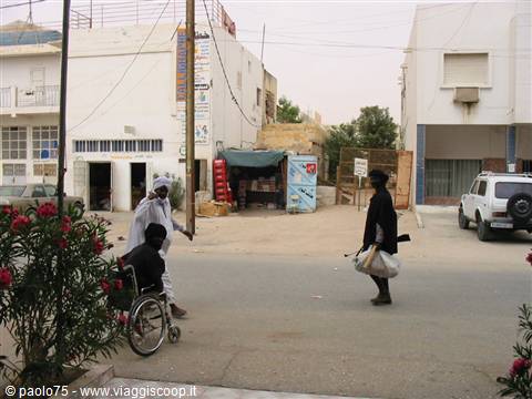 man pushing wheelchair and veiled woman walking along street between white buildings