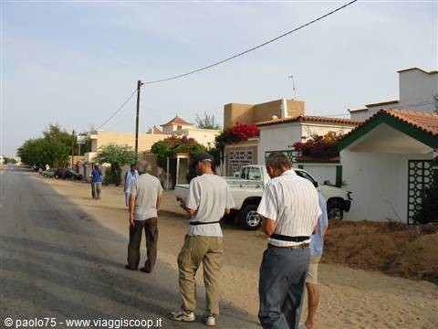 Tourists walk along sandy road past white houses with red flowers