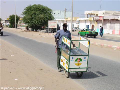 Vendor pushes painted ice cream cart along sandy roadside under harsh sun