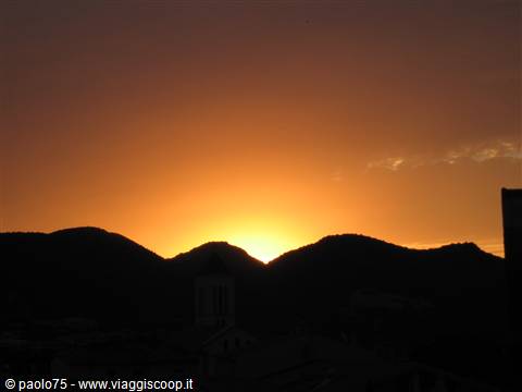 Sun glowing between dark hills, a bell tower and rooftops silhouetted