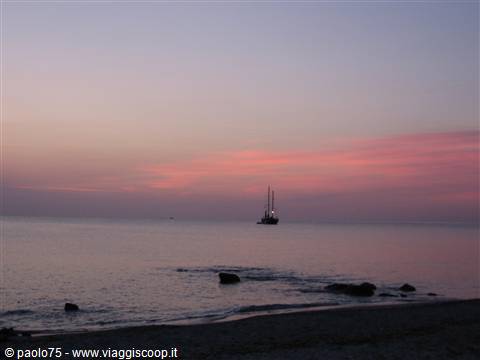 Sailboat silhouetted on calm sea at dawn, rocks on the beach