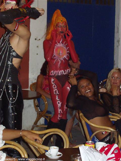 Dancer painted in red with orange wig performs between café tables at night