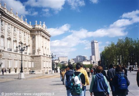 Tourists with backpacks walk toward the Royal Palace of Madrid under cloudy skies