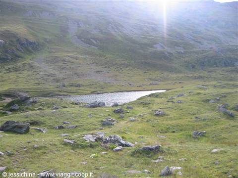 Alpine tarn among green meadows and rocks, bright sun over mountains