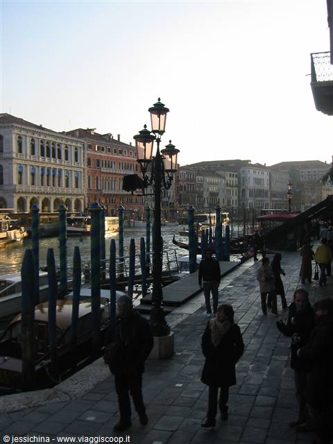 Pedestrians on the stone quay in Venice, historic palaces and lit lampposts along the Grand Canal