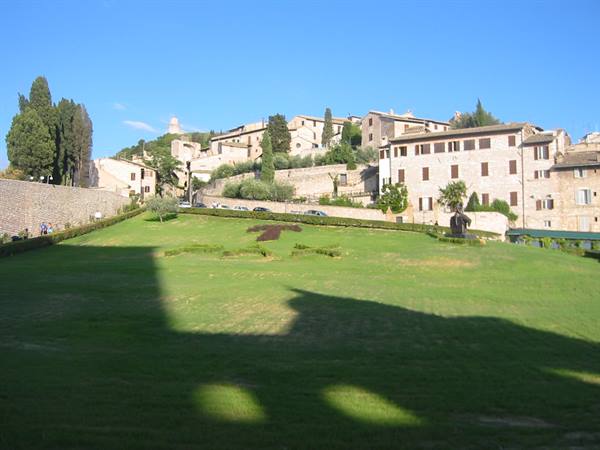 Green lawn with stone shadows in front of Assisi houses under blue sky