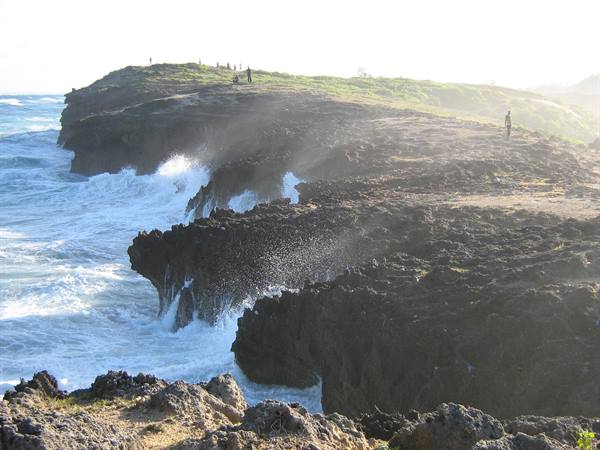 Tourists on rocky cliff above crashing waves, intense sunlight