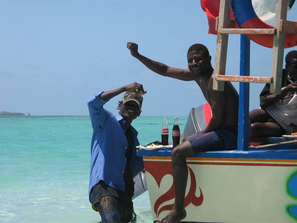 Two men waving beside a painted wooden boat in turquoise water
