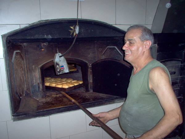 Baker smiles beside wood-fired oven with freshly baked bread