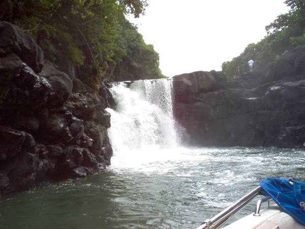 Waterfall over dark rocks seen from a boat, lush trees