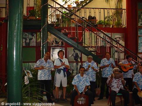 Musicians in floral shirts play percussion and guitar beneath an iron staircase