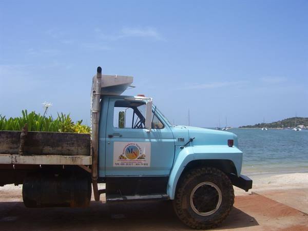 Blue truck loaded with green plants, parked on the beach near boats