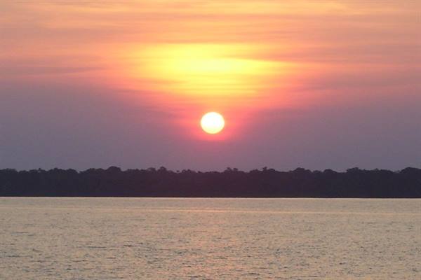 Orange sun low over dark treeline, pink light on the water