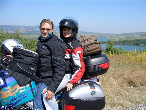 Two motorcyclists in gear beside their touring bike, luggage cases, lake and green hills