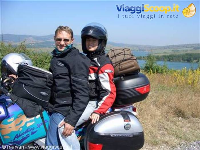 Two motorcyclists in gear beside their touring bike, luggage cases, lake and green hills