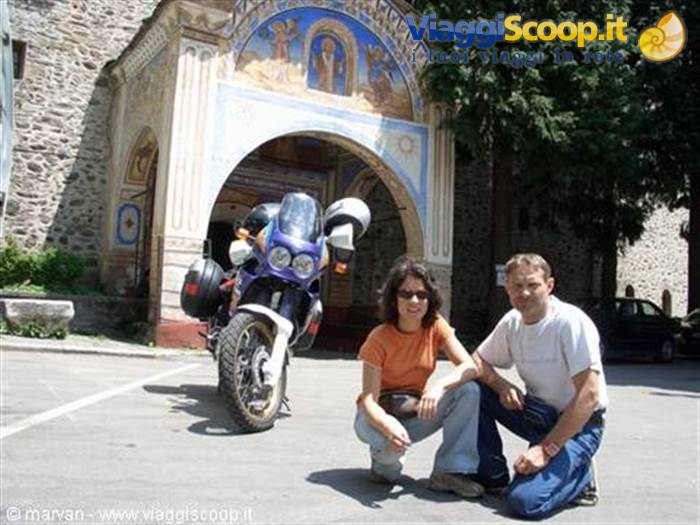 Couple crouching in front of adventure motorcycle under the frescoed arch of a stone monastery