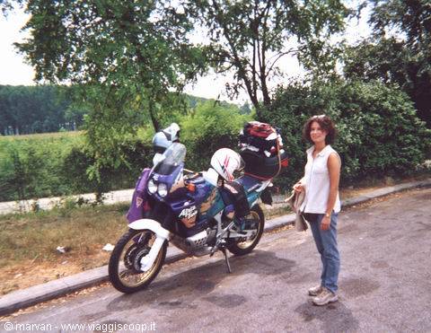 Woman standing beside a loaded touring motorcycle on a tree-lined country road