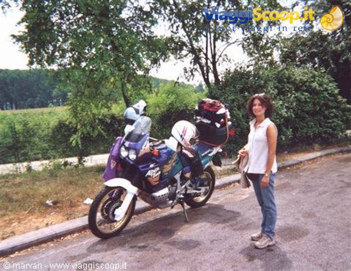 Woman standing beside a loaded touring motorcycle on a tree-lined country road